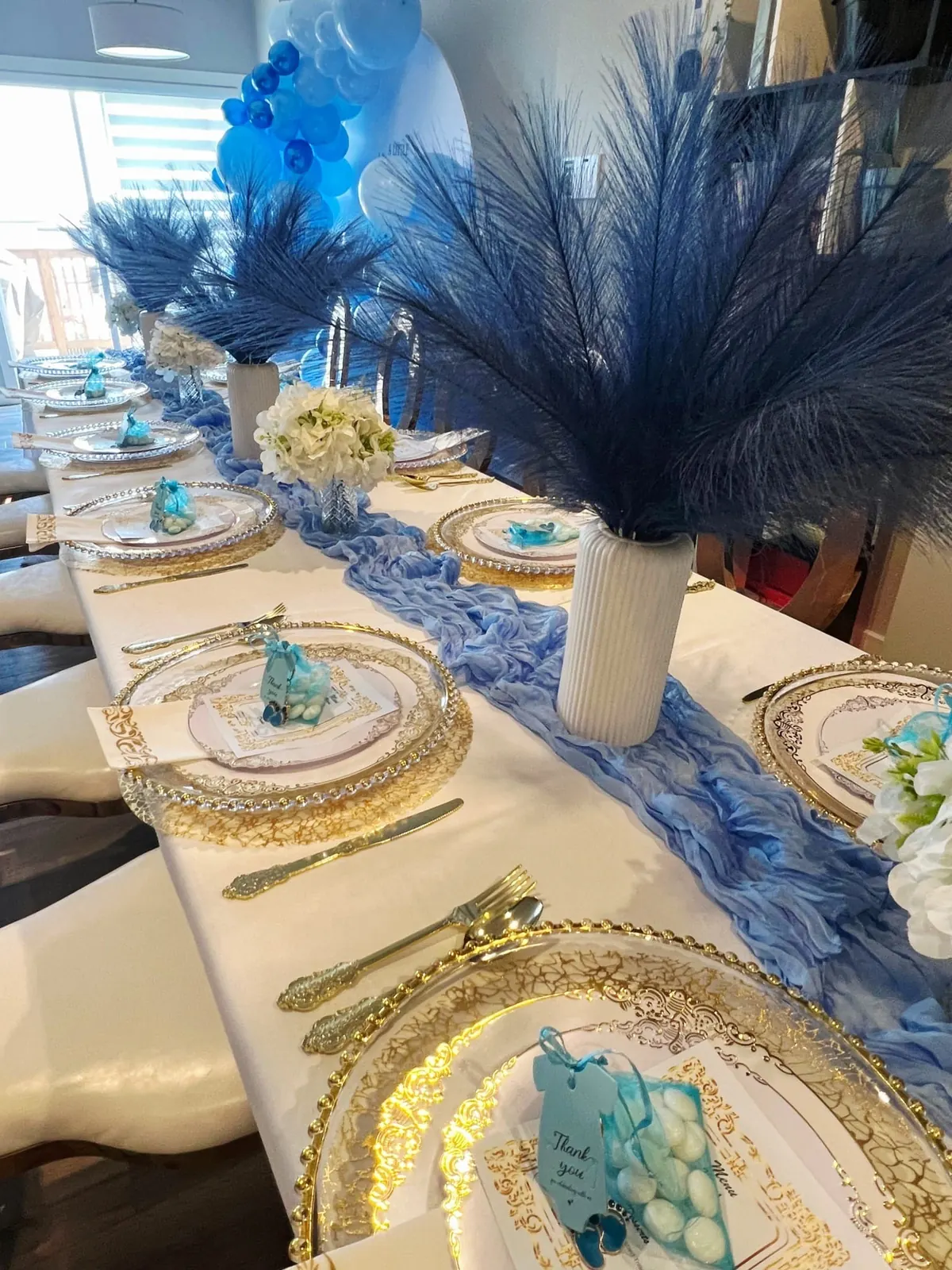 A baby shower table with a blue runner, gold chargers and pampas stems